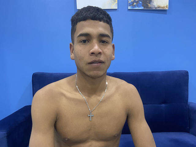 JhonBennet is shirtless with a slim, toned chest, brown skin, short black hair, and a silver cross necklace. He's sitting on a blue sofa, looking at the camera.