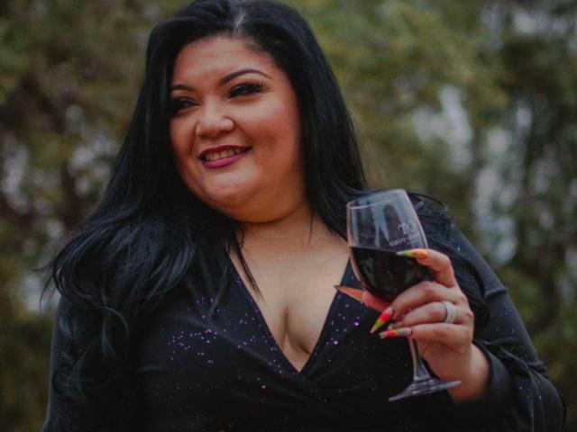 LaCurvy, com cabelo preto, segurando um copo de vinho tinto, sorrindo, vestindo um vestido preto brilhante, exibindo seus seios grandes e usando unhas coloridas, em um cenário de floresta..