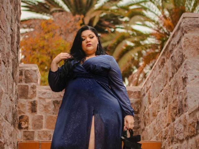LaCurvy with big ass, wearing a shiny blue dress with a thigh-high slit, black hair, holding a black bag, standing on stone steps, touching her hair.