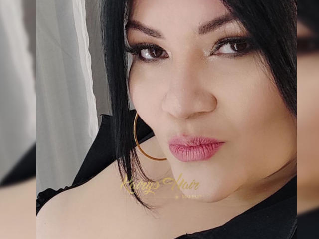 LaCurvy tem a pele marrom média, cabelo preto e está usando batom rosa com um brinco de argola. Seus seios estão ligeiramente visíveis sob uma blusa preta..