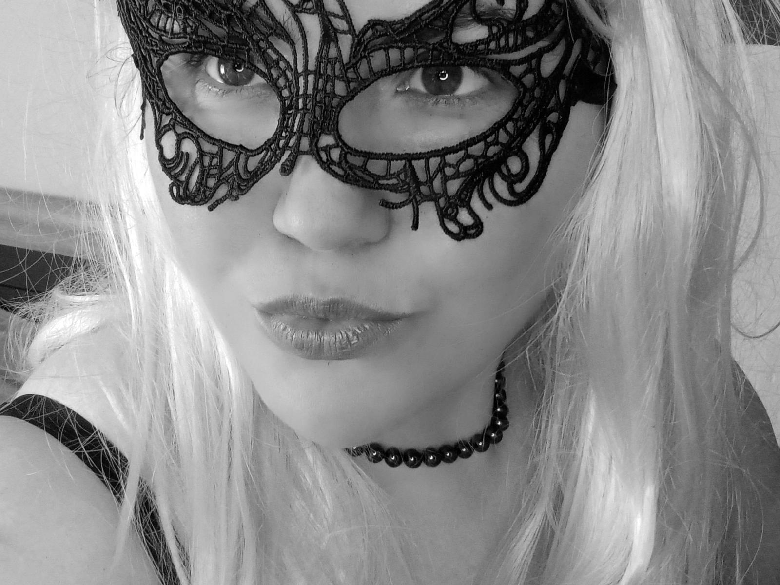 Photo en noir et blanc de MilfVanessaJones avec de longs cheveux blonds, portant un masque en dentelle complexe, un collier de perles et un rouge à lèvres brillant, regardant l'appareil photo avec un sourire discret..