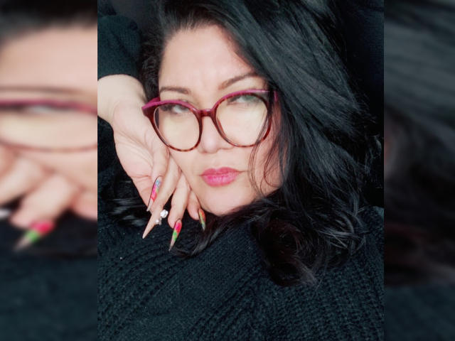 LaCurvy has dark wavy hair, red glasses, and pink lipstick. She's wearing a black knit sweater with colorful nails and a ring on her right hand.