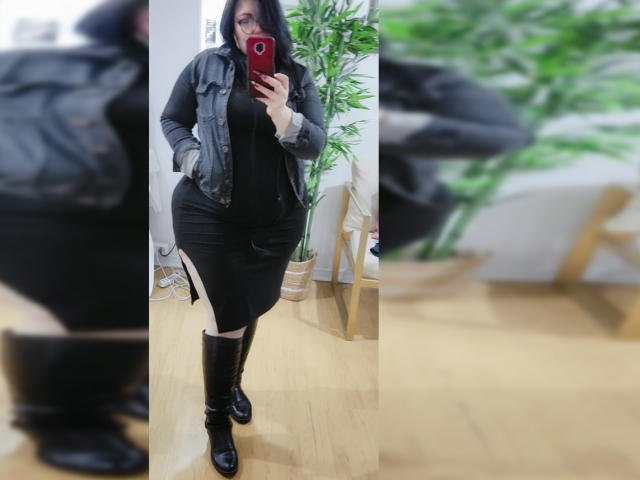 LaCurvy vestindo um vestido preto, jaqueta cinza e botas pretas até o joelho, segurando um telefone vermelho, em pé, exibindo suas curvas e seios grandes, com uma planta verde ao fundo, e piso de madeira..