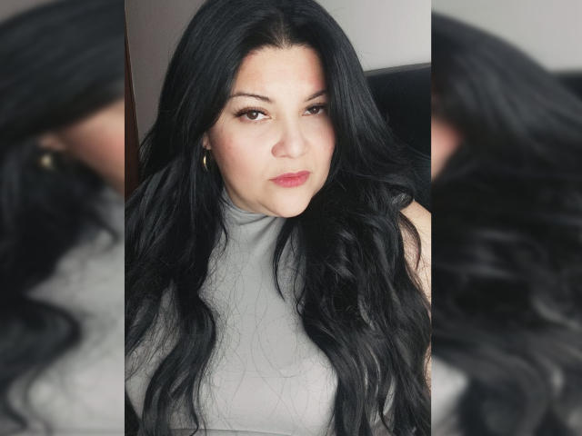 LaCurvy with long black hair, wearing a grey turtleneck, has a serious expression, subtle red lipstick, and hoop earrings, with a slight blush on her right cheek.