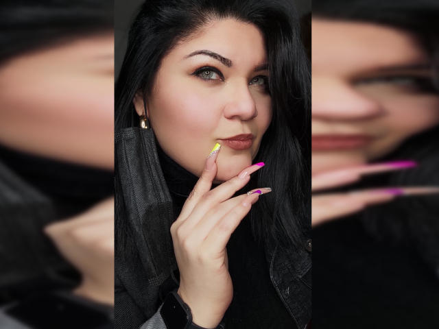 LaCurvy with black hair, green eyes, and pink-yellow nails, touching her lip with a finger. Blurred background of her face. Sheer black jacket. LaCurvy's tits are slightly visible.