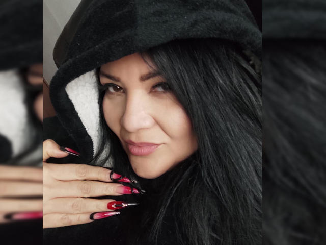 LaCurvy with long black hair, wearing a black hood, smiling with pink lips, black nails with red tips, touching her face, looking at camera.