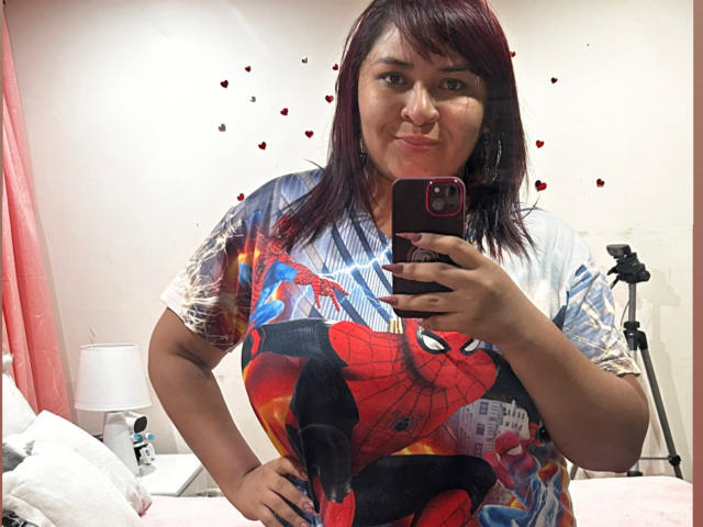KarlaWild, avec ses cheveux roux foncé, sa poitrine de taille moyenne, portant un t-shirt Spider-Man, tenant un téléphone rouge, prenant un selfie devant un miroir, souriant légèrement, debout dans une chambre..