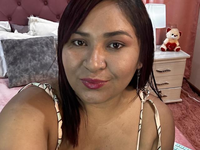 KarlaWild, a chubby Latina woman with dark brown hair, wearing a patterned top, pink lipstick, nose piercing, and earrings, sitting in a pink room. Boobs visible.
