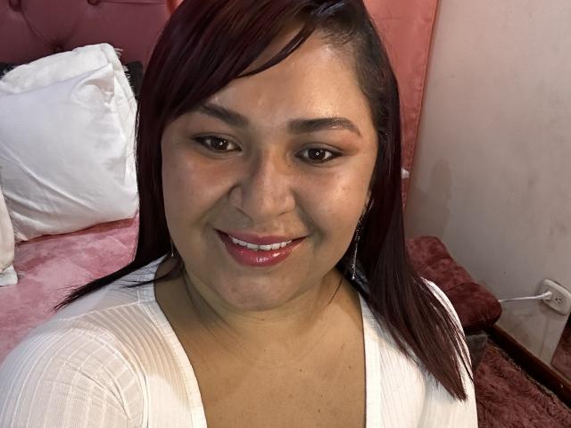 KarlaWild with medium brown skin, red lipstick, and straight dark brown hair, smiling at the camera. She's wearing a white top and has small, round tits.