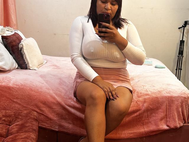 KarlaWild, with tan skin and dark brown hair, sits on a pink bed, wearing a white ribbed top and pink skirt, taking a selfie with her phone, showing her tits.
