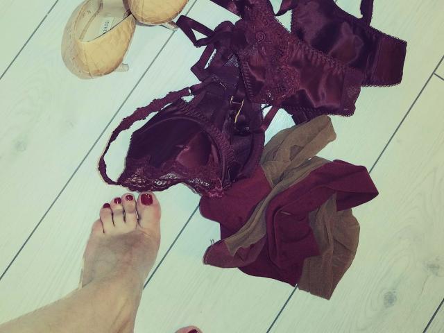 WandaDominatrix's feet with red nail polish, above a pile of maroon lace lingerie and brown pantyhose on white wooden floor, with beige high heels in the top left.