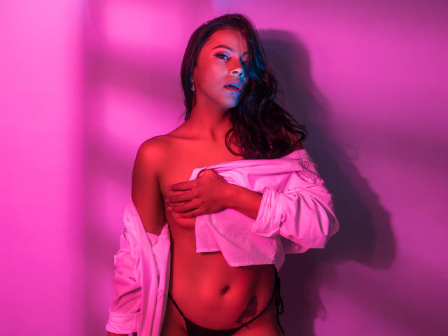 Amber, con i capelli scuri, illuminata di rosa, che copre il seno con una camicia bianca, indossa uno slip nero e ha l'ombretto blu, e guarda seducentemente la fotocamera..