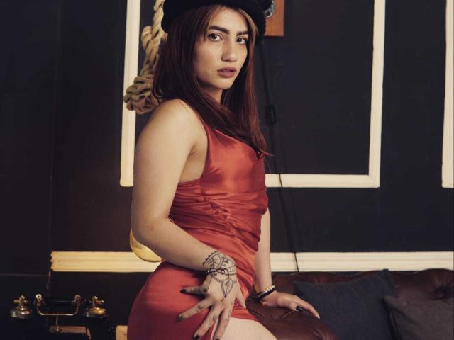 SophiaHottie con vestido rojo, sombrero negro, tatuaje en la muñeca, cabello castaño, mirando a la cámara, sentada en un sofá de cuero marrón, con la mano en el trasero, fondo oscuro, cuerda visible..