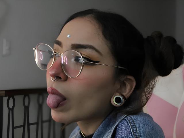 LemoonPeach porte des lunettes, un anneau de nez et des élargisseurs d'oreilles, avec un troisième œil sur son front, la langue tirée, et une veste en jean bleu. Ses seins ne sont pas visibles..
