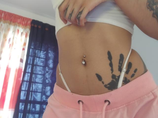 La sezione centrale di LemoonPeach, che mostra un ombelico con piercing, un tatuaggio a forma di mano nera sul lato destro, pantaloncini rosa con spalline bianche e una canotta bianca con tatuaggi sulla mano..