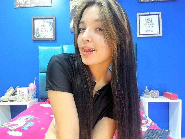 SarahGood with long brown hair, black t-shirt, and tongue out, leaning on a bed with pink fairy-themed sheets, blue wall in background, and white shelves with shoes.