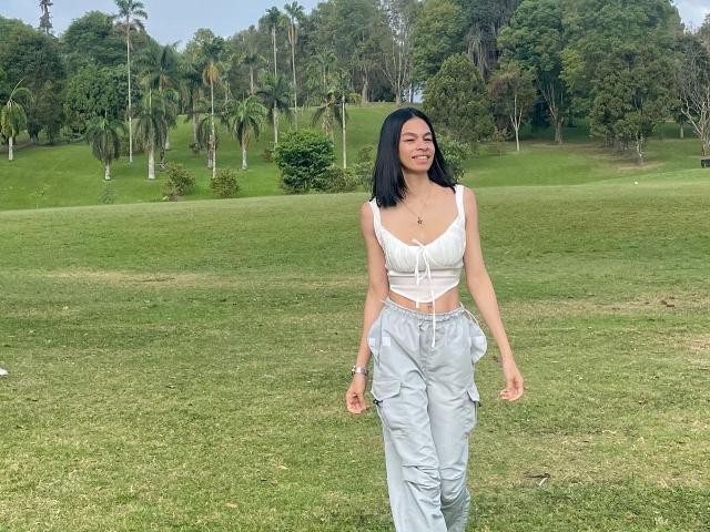 AmberGray69 has small tits, wearing a white crop top and light gray pants, with black hair, smiling, standing on a grassy field with palm trees in the background.