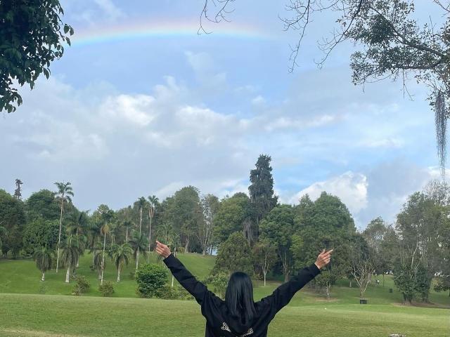AmberGray69 stands with arms raised, back to camera, wearing a black hoodie, in a grassy park with trees and a rainbow in the cloudy sky.