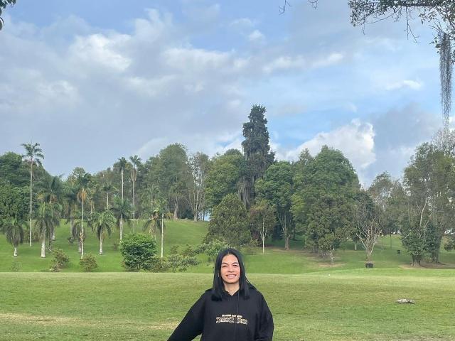 AmberGray69 con sudadera negra, de pie en hierba verde, sonriendo, cabello negro de longitud media, piel pálida, senos pequeños, brazos ligeramente levantados, palmeras y cielo nublado en el fondo..