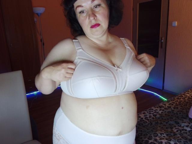 ErinSmart, une femme corpulente avec des cheveux noirs courts, portant un soutien-gorge blanc et des sous-vêtements assortis, tire sur son soutien-gorge pour montrer ses seins volumineux et pâles dans une chambre..