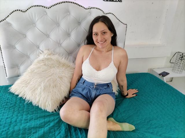 AnhaliaSkarleth sits on a green quilted bed, wearing a white lace top and denim shorts, with a fluffy pillow, smiling at the camera, showing her tits and legs.