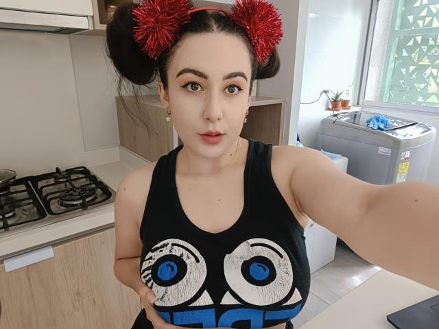MabyLegrand with puffy red hair buns, wearing a black tank top with big eye design, showing off her sizable boobs in a kitchen, taking a selfie.