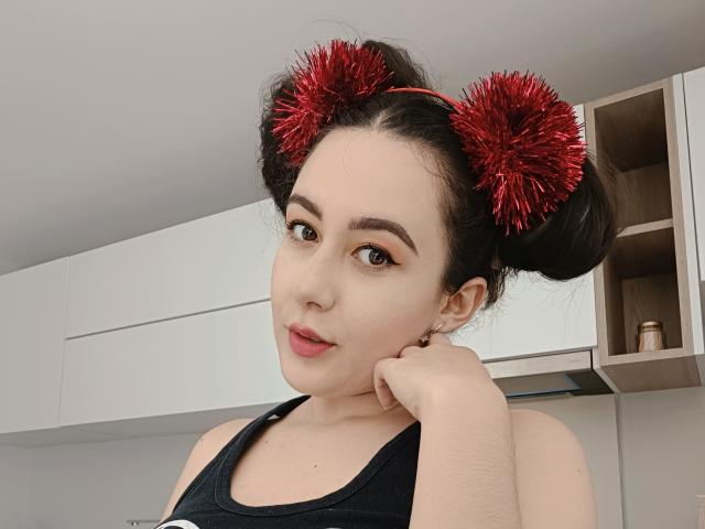 MabyLegrand with red fuzzy pom-pom hair buns, black tank top, light skin, brown eyes, pink lips, hand on cheek, in a modern kitchen. Her tits are visible.