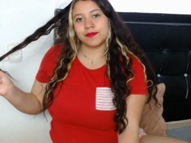 KrisGarner has long, wavy black and blonde hair, red shirt, nose ring, and red lipstick. She's holding her hair with a cute smile, showing her tits.