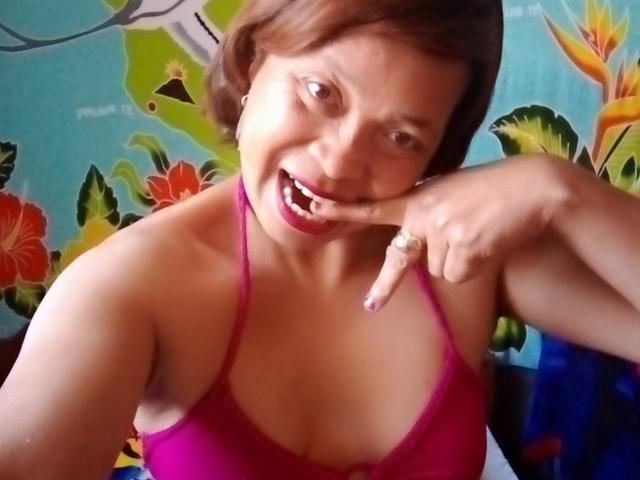 Rahel69, with short brown hair, smiling and biting her finger, wears a pink halter top, showing her tits and a ring on her finger, against a colorful floral background.