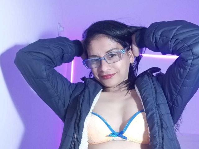 MatureEstery met een blauwe bril, zwart haar en rode lippenstift, draagt een blauwe gewatteerde jas en een gele bh met blauwe randen, haar handen in haar haar, lachend. Borsten zichtbaar..