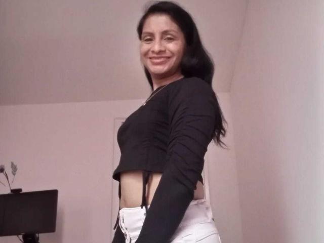 Eine reife Frau namens Estery mit langen, schwarzen Haaren, die lächelt, trägt ein schwarzes, langärmeliges Crop-Top und weiße, hochgeschnittene Hosen. Dabei sind ihr nackter Bauch und schwarze Träger ihrer Unterwäsche zu sehen..