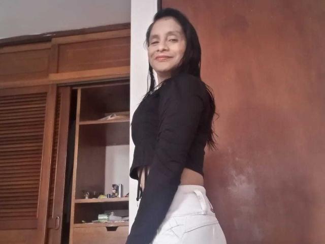 MatureEstery with long black hair, wearing a black top and white pants, smiling at the camera, standing sideways, showing a bit of her white pants' ass.