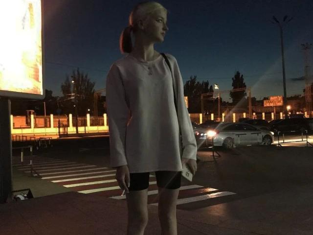 LaPochka, met een kaal hoofd, draagt een losse roze trui en zwarte korte broek, staat op een zebrapad 's nachts, met een telefoon in haar hand, met straatverlichting en auto's op de achtergrond..