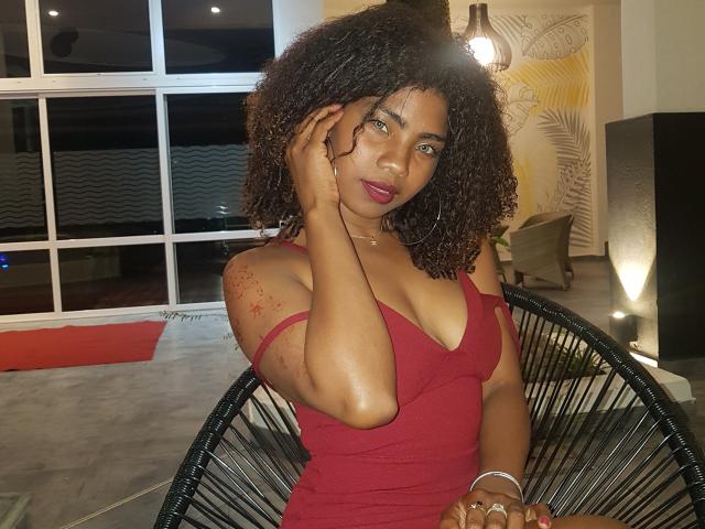 RonaSweet, avec ses cheveux bruns bouclés, une robe rouge, montrant sa poitrine généreuse, une main sur son visage, du rouge à lèvres rouge foncé, assise sur une chaise noire, dans une pièce éclairée par une lumière chaude..
