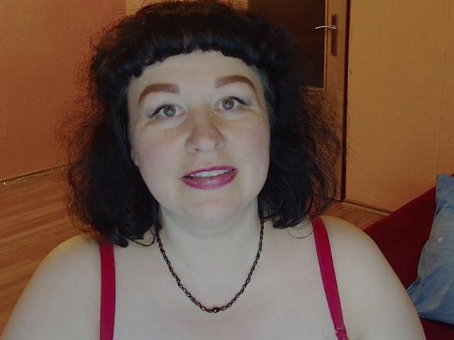 ErinSmart tem cabelo preto encaracolado, pele clara e está vestindo uma camiseta vermelha com um colar de corrente preta. Ela está ligeiramente de língua para fora. Seios visíveis..