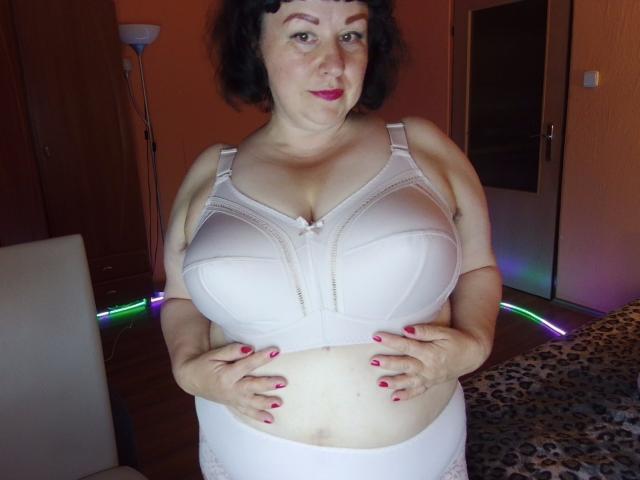 ErinSmart, eine kurvige Frau mit großen Brüsten, trägt einen weißen BH und hält ihren Bauch, hat kurze schwarze Haare und roten Lippenstift und steht in einem Raum mit LED-Leuchten..