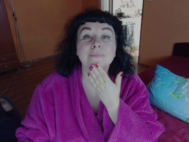 ErinSmart with curly black hair, wearing a pink bathrobe, has red nails and is touching her neck, smiling with blue eyes. Her tits are partially visible under the robe.