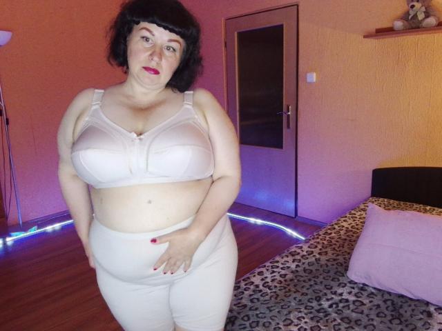 ErinSmart has large tits in a sheer white bra, plus-size with a round belly, wearing matching white pants, standing in a bedroom with a leopard-print bed.