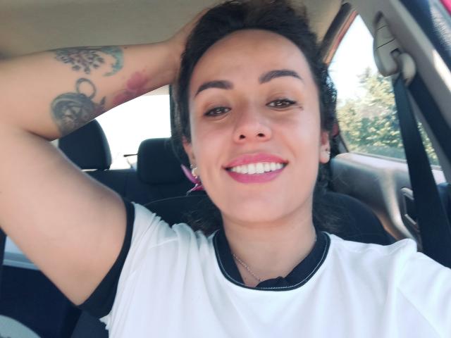 Joanallen, sorrindo com batom rosa, tem cabelo escuro, olhos castanhos e uma tatuagem no braço direito, vestindo uma camisa branca com mangas pretas, sentada em um carro..