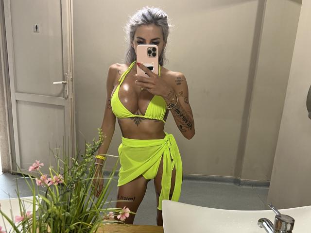 LisaaKiraa, avec un haut de bikini vert brillant et un paréo assorti, montrant ses seins et un tatouage sur son bras droit, prend un selfie devant un miroir dans une salle de bain..