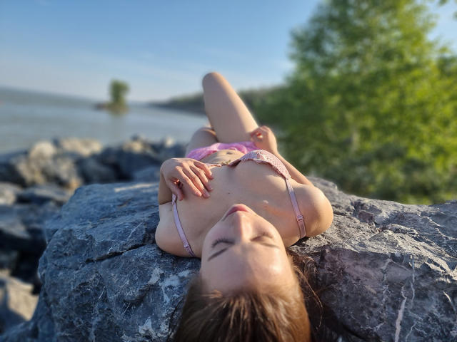 KarlyLeen, con senos pequeños y un sujetador rosa, está acostada sobre una roca, vistiendo ropa interior rosa, con los brazos por encima de la cabeza, los ojos cerrados y la luz del sol sobre su piel. En el fondo, hay un árbol verde..