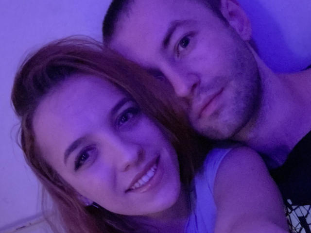 EvaMaks, avec ses courts cheveux bruns et son eye-liner noir, souriante, se penche sur un homme barbu et avec des poils de barbe, tous deux sous un éclairage violet. Le visage de l'homme est légèrement flou..