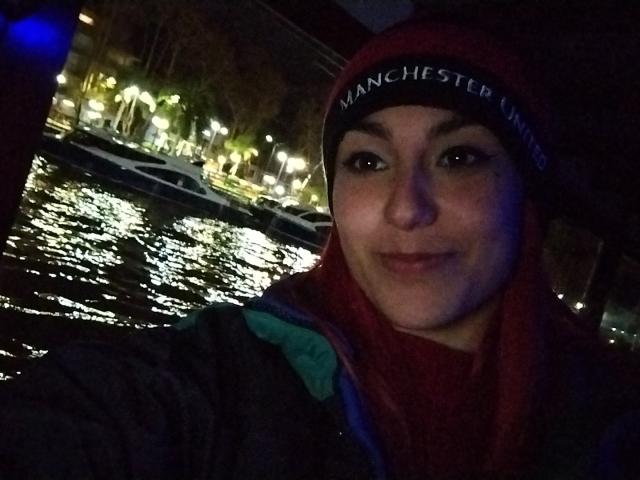 AmyStreetwise69 hat ein süßes Lächeln, dunkle Augen und ein rotes Kopftuch, trägt eine schwarze Mütze und eine grüne Jacke und steht nachts an einem dunklen, reflektierenden Gewässer..