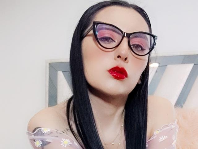 NataliaCamp, com cabelo preto, óculos, maquiagem rosa nos olhos, batom vermelho e uma blusa transparente com estampa de margaridas, olhando ligeiramente para o lado com os lábios franzidos..