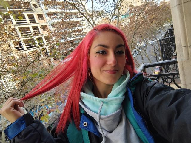 AmyStreetwise69 with bright red hair, holding a strand, wearing a green jacket over a blue hoodie, smiling, blue eyes, light skin, outdoors with trees in the background.