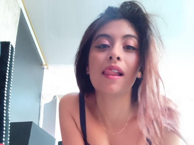 SamanthaaSmith with pink-tipped black hair, black strap top, tongue out, pink eyeliner, earrings, necklace, and a slight smile, looking down at the camera.