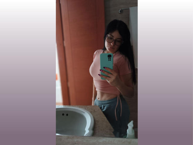 VaaleryStark with glasses, pink top, grey pants, taking a mirror selfie with a teal phone, showing her small tits and slim ass, in a bathroom with a sink.