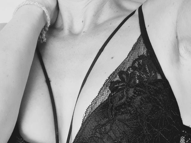 Les seins d'ApoloniaRed, dans un soutien-gorge noir en dentelle, avec des bretelles fines, un décolleté visible, portant un bracelet délicat à son bras droit, en noir et blanc, gros plan, éclairage doux..
