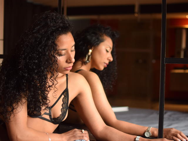 Jan Evans, avec ses cheveux noirs bouclés, portant un soutien-gorge en dentelle noire, est appuyée contre un miroir, reflétant son profil, montrant ses fesses et ses petites seins, dans une pièce faiblement éclairée..