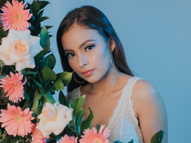 KiaraFire mit langen, braunen Haaren, die ein weißes Spitzenkleid, goldene Ohrringe im Stecker-Stil und rosa Rosen trägt, hat ein sanftes Lächeln und dezentes Make-up und steht neben einer Blumenarrangements..